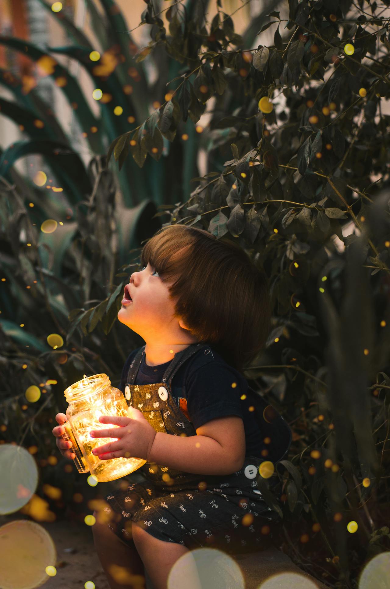 A young boy with brown hair holds a jar of glowing fireflies. He sits in front of a tree. He is very calm, and not anxious.